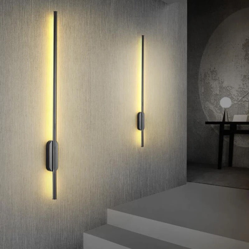 Long aluminum stick-shaped modern wall light in black finish illuminating the wall with soft warm white glow