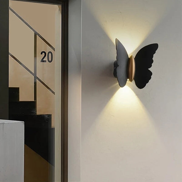 A black outdoor wall light in the shape of a butterfly mounted on the wall, emitting a warm white glow that casts a soft, ambient light around the area