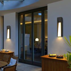 Modern outdoor LED wall light mounted on a house's exterior wall, emitting a warm white glow, highlighting its sleek design and illuminating the surrounding area.