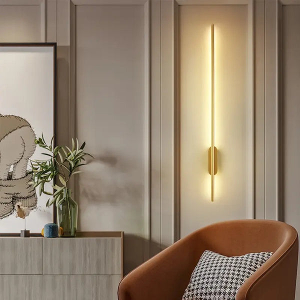 Long stick-shaped modern wall light in gold finish illuminating the wall with soft warm white glow
