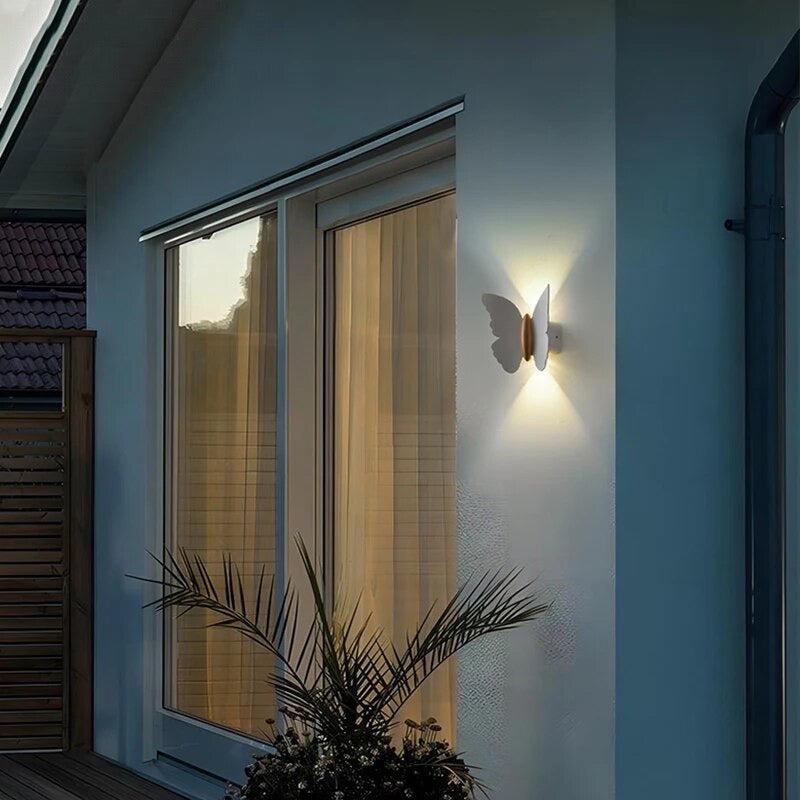 A white outdoor wall light in the shape of a butterfly mounted on the wall, emitting a warm white glow that casts a soft, ambient light around the area